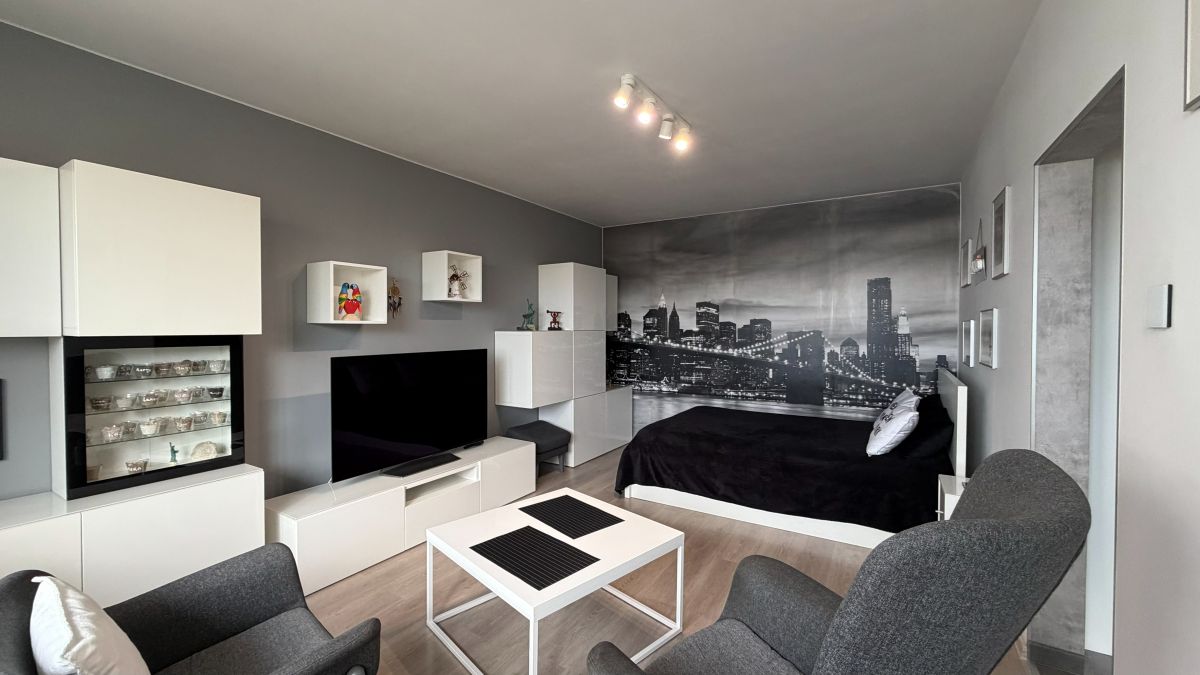 Modern studio with white modular furniture, gray walls, and a black bed against a city skyline mural at the far wall.