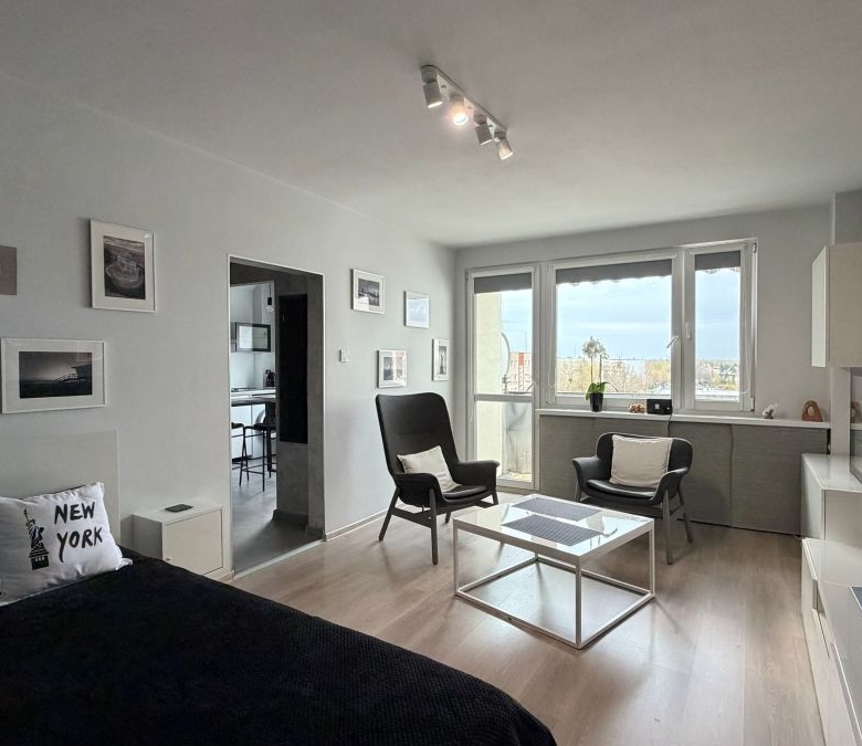 Bright modern studio living area with a bed on the left, black bedding and New York pillows, two black armchairs, a white glass coffee table, and large windows.