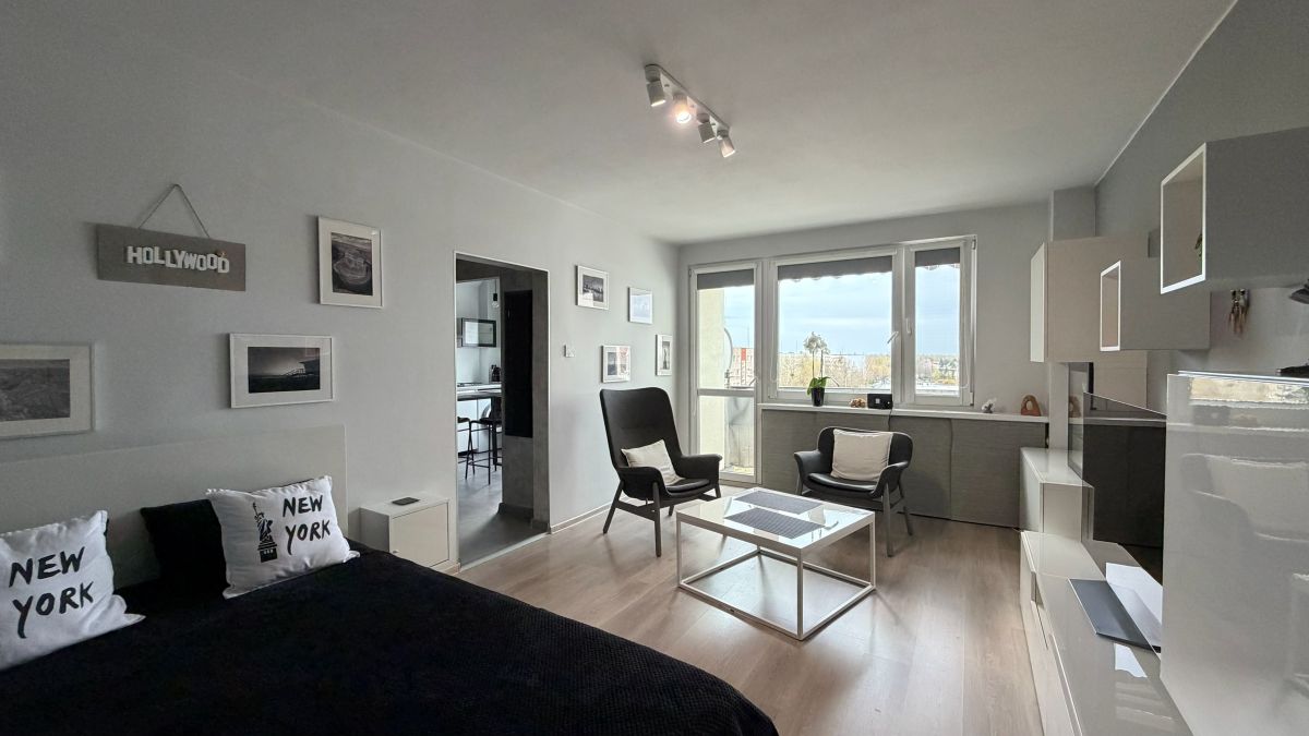 Bright modern studio living area with a bed on the left, black bedding and New York pillows, two black armchairs, a white glass coffee table, and large windows.