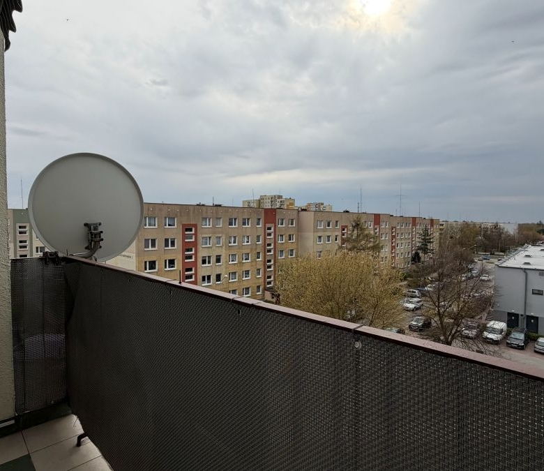 Balcony with a satellite dish overlooking a row of apartment buildings and a parking lot under a cloudy sky