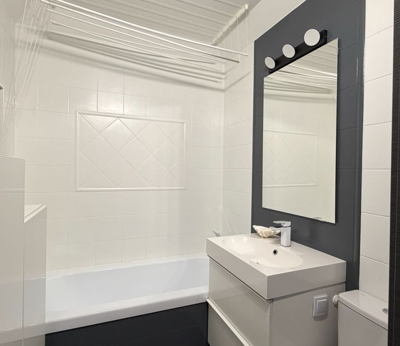 Small modern bathroom with white tiled walls, dark accent wall, rectangular mirror, white sink vanity, toilet, and bathtub.