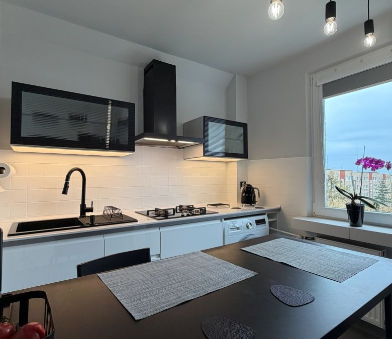Modern white kitchen with black accents, gas stove, and hanging pendant lights; a dark dining table with placemats in the foreground.