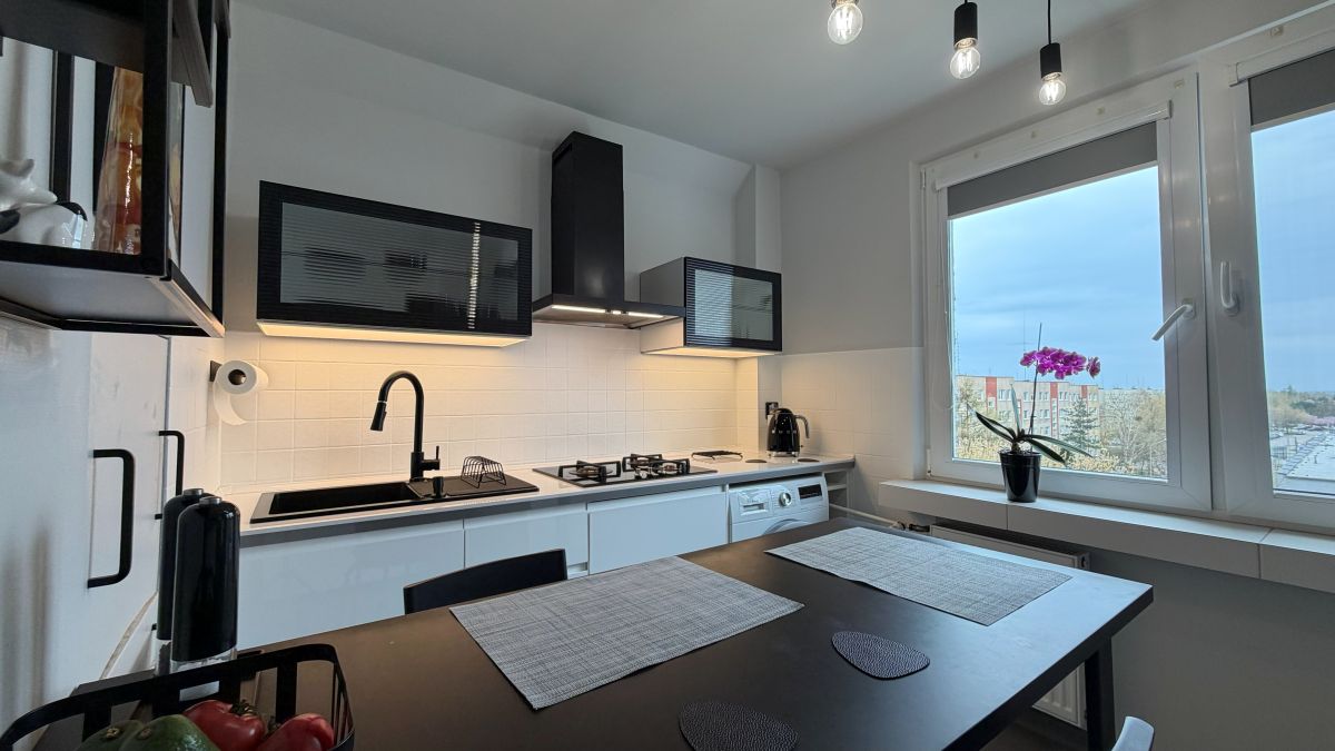 Modern white kitchen with black accents, gas stove, and hanging pendant lights; a dark dining table with placemats in the foreground.