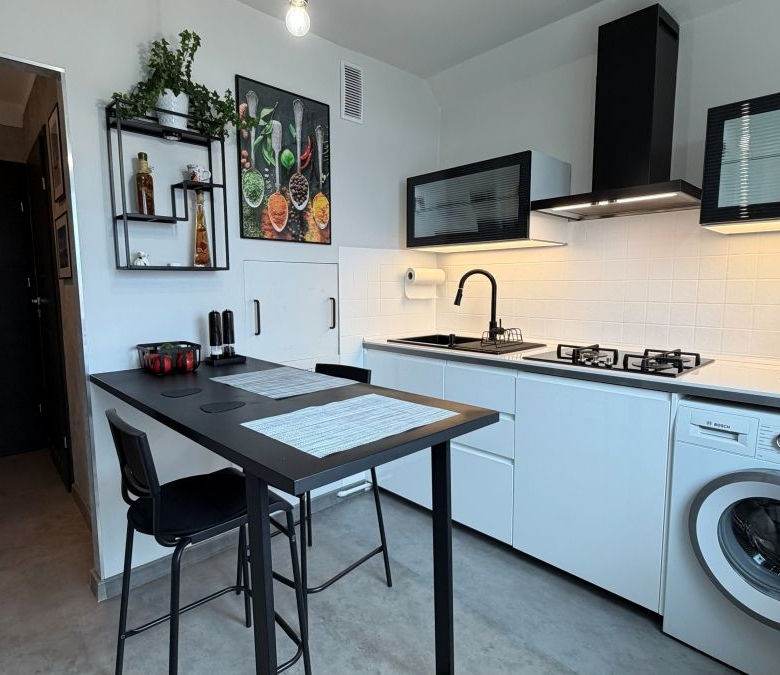 Modern white kitchen with a black breakfast bar, two black stools, and a washing machine under the counter; features a stove, kettle, and hanging range hood.
