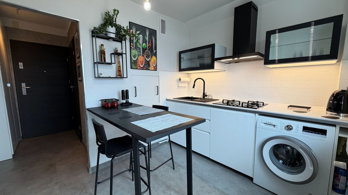 Modern white kitchen with a black breakfast bar, two black stools, and a washing machine under the counter; features a stove, kettle, and hanging range hood.