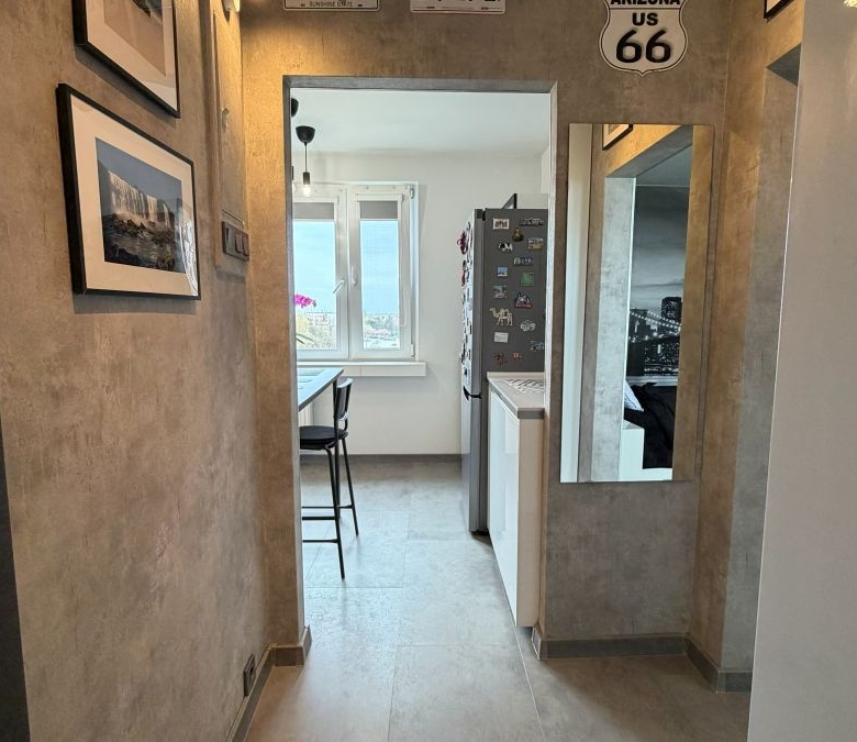Narrow hallway leading to a small kitchen with a window, fridge magnets, and a bar stool at a counter.