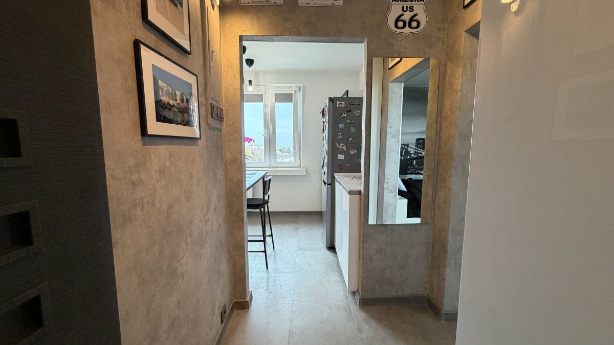 Narrow hallway leading to a small kitchen with a window, fridge magnets, and a bar stool at a counter.