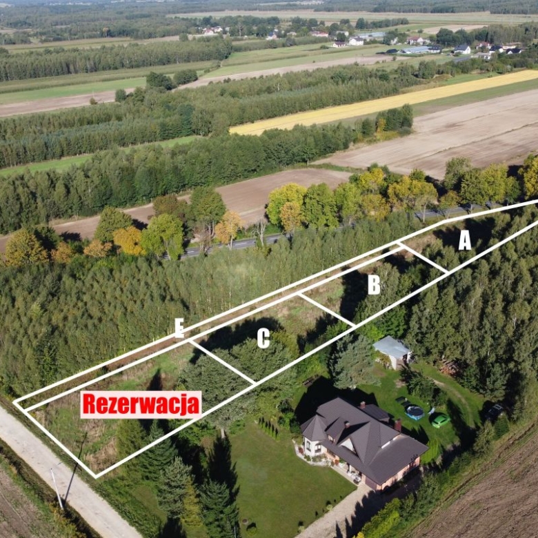 Aerial view of a rural property with a long triangular lot outlined in white and divided into sections labeled A through F along a tree-lined road; a red 'Rezerwacja' sign marks the first parcel.