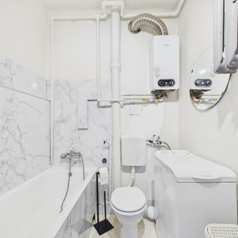 Small white utility bathroom with bathtub, toilet, washing machine, and exposed pipes; wall-mounted boiler visible above.
