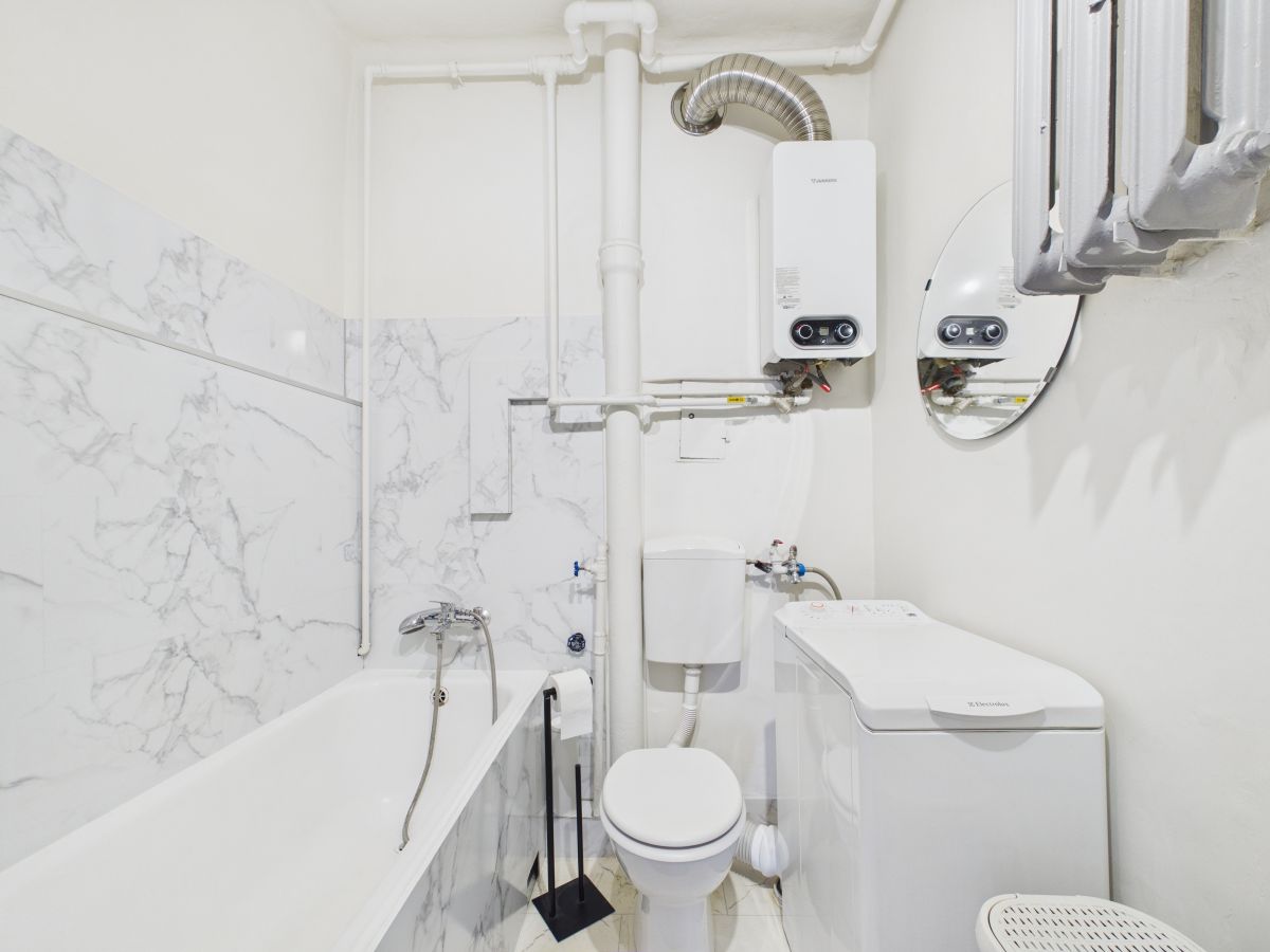 Small white utility bathroom with bathtub, toilet, washing machine, and exposed pipes; wall-mounted boiler visible above.