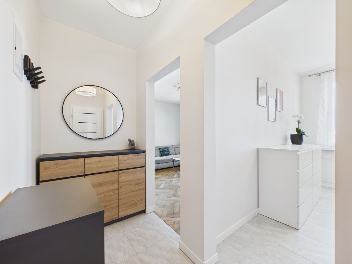 Hallway with a wooden and black cabinet under a round mirror, coat hooks on the left, leading to a living room with a gray sofa seen through the doorway.