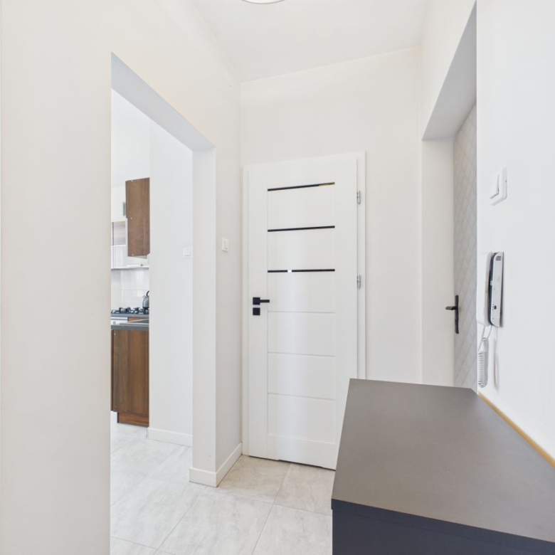 Bright white hallway leading to a white panel door, with an intercom on the right wall and a dark countertop along the side.