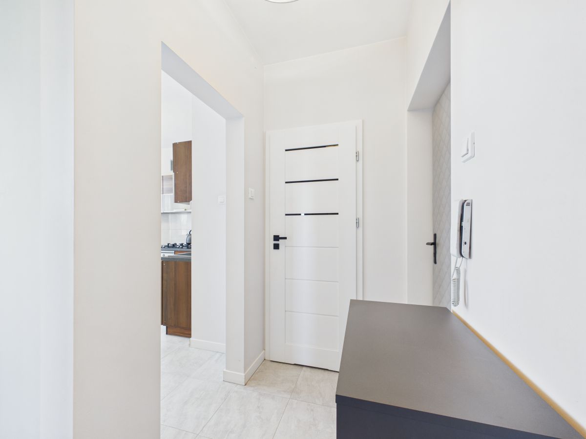 Bright white hallway leading to a white panel door, with an intercom on the right wall and a dark countertop along the side.