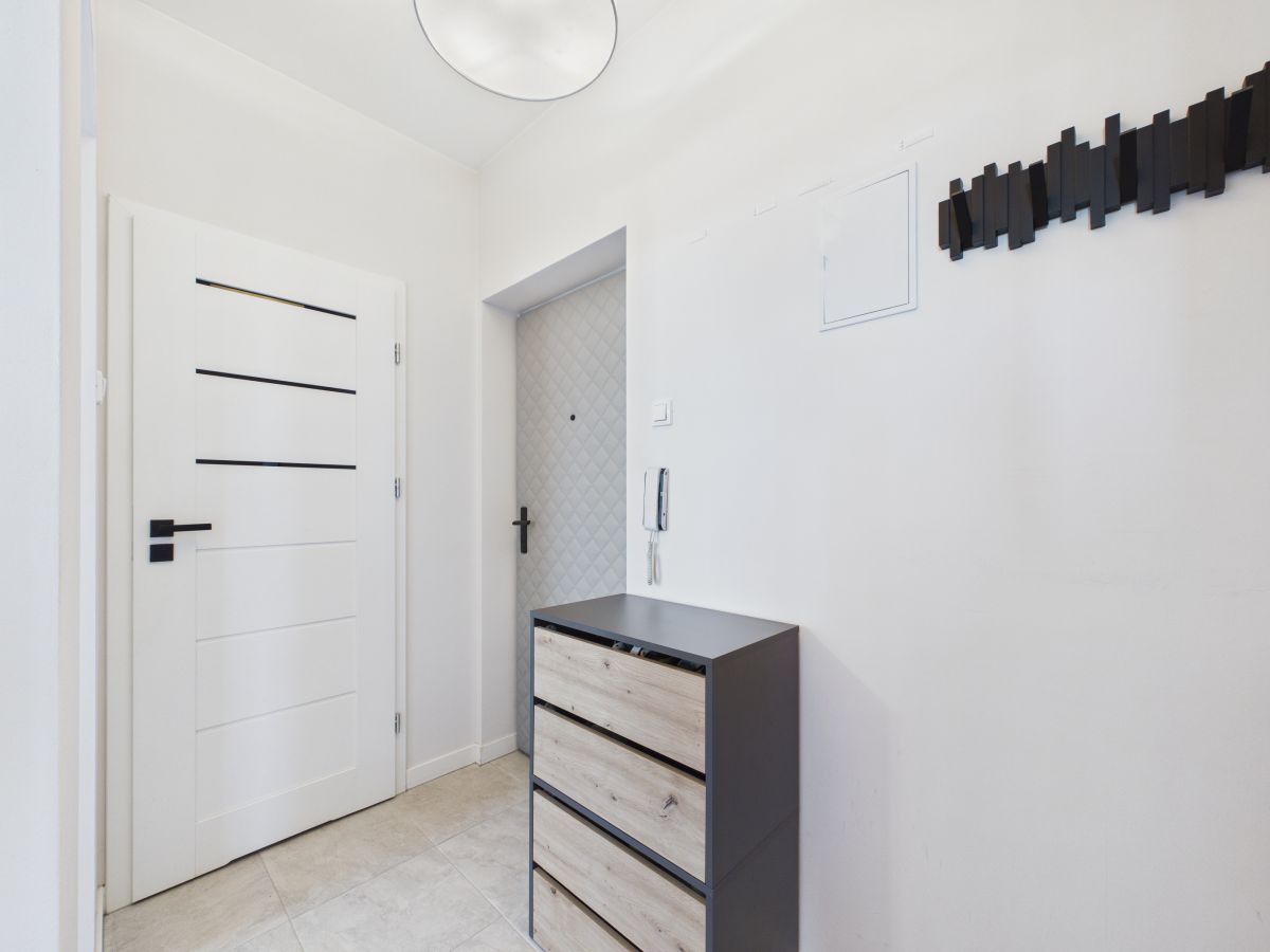 Minimalist white-entry hallway with a white paneled door, a gray dresser with light wood drawers, and a black wall coat rack on the right.