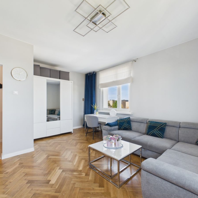 Bright modern living room with a gray L-shaped sofa, white storage cabinet, and a glass-top coffee table on a herringbone wood floor.
