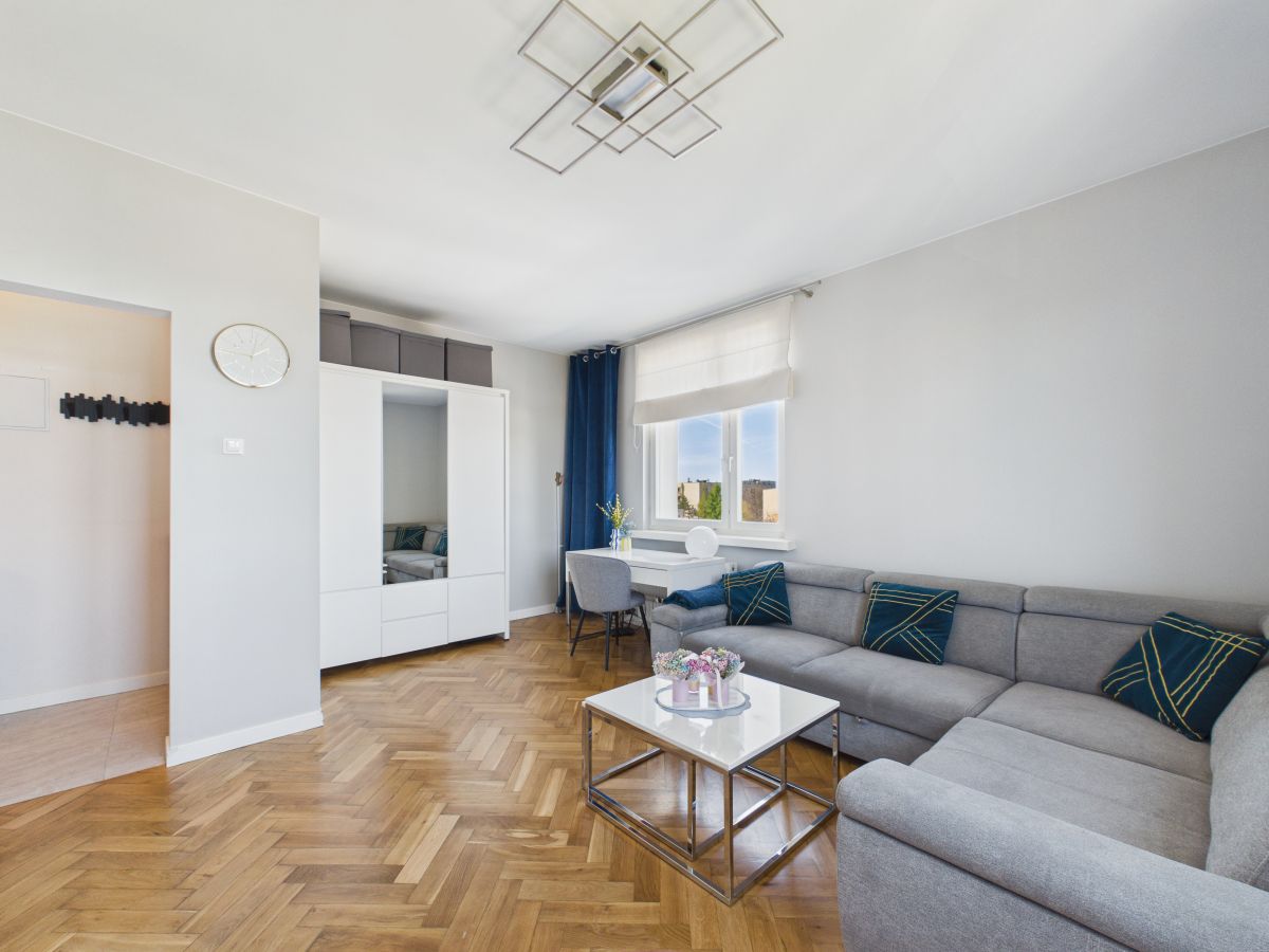 Bright modern living room with a gray L-shaped sofa, white storage cabinet, and a glass-top coffee table on a herringbone wood floor.