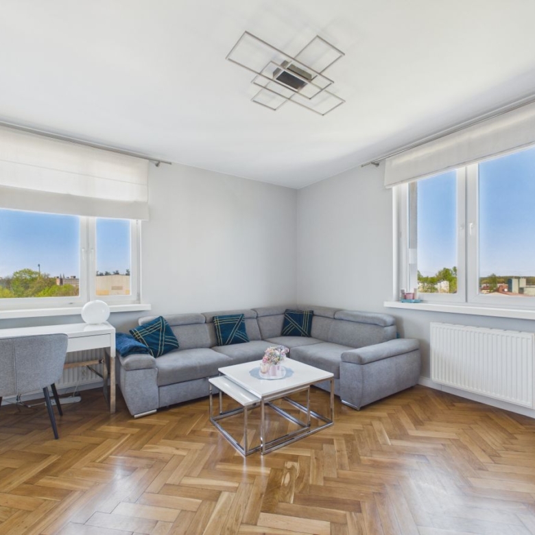 Bright living room with a gray L-shaped sofa, white coffee table, and two large windows with white blinds and blue curtains.