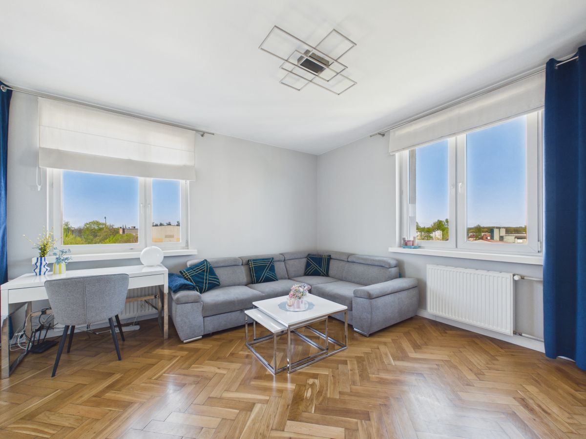 Bright living room with a gray L-shaped sofa, white coffee table, and two large windows with white blinds and blue curtains.