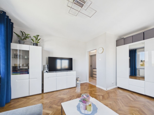Bright minimalist living room with white storage cabinets, a flat-screen TV on a white media console, and blue curtains over a herringbone wooden floor.