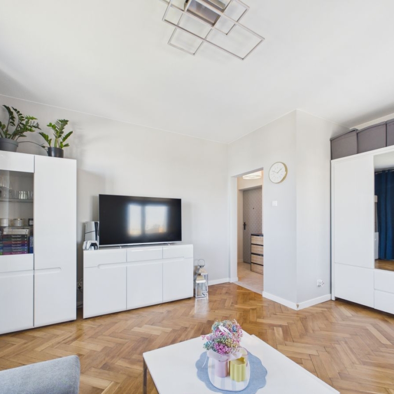 Bright minimalist living room with white storage cabinets, a flat-screen TV on a white media console, and blue curtains over a herringbone wooden floor.
