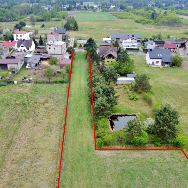 Aerial view of a long, L-shaped rural plot outlined in red, featuring a central grassy lawn, a small pond, and trees near houses.