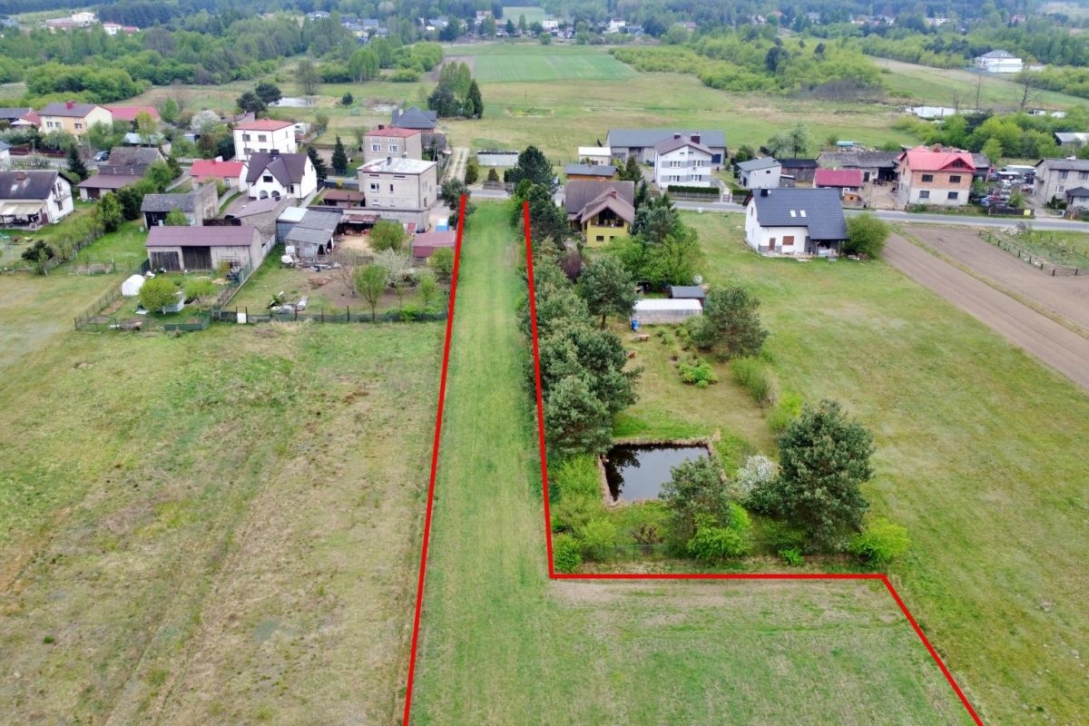 Aerial view of a long, L-shaped rural plot outlined in red, featuring a central grassy lawn, a small pond, and trees near houses.