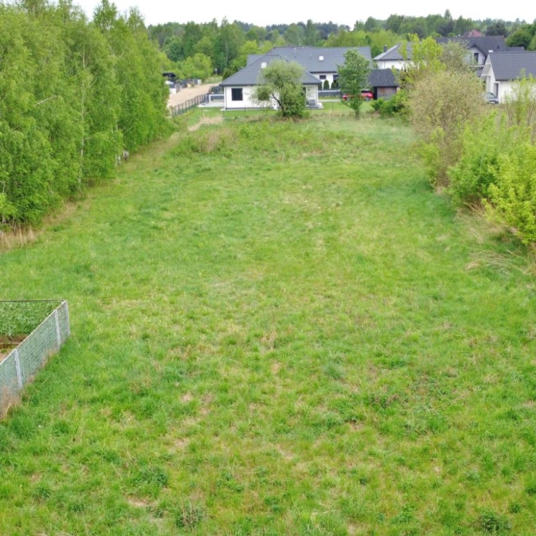 Aerial view of a grassy open plot bordered by trees on both sides, with houses visible in the distance and a fenced garden area in the lower left.