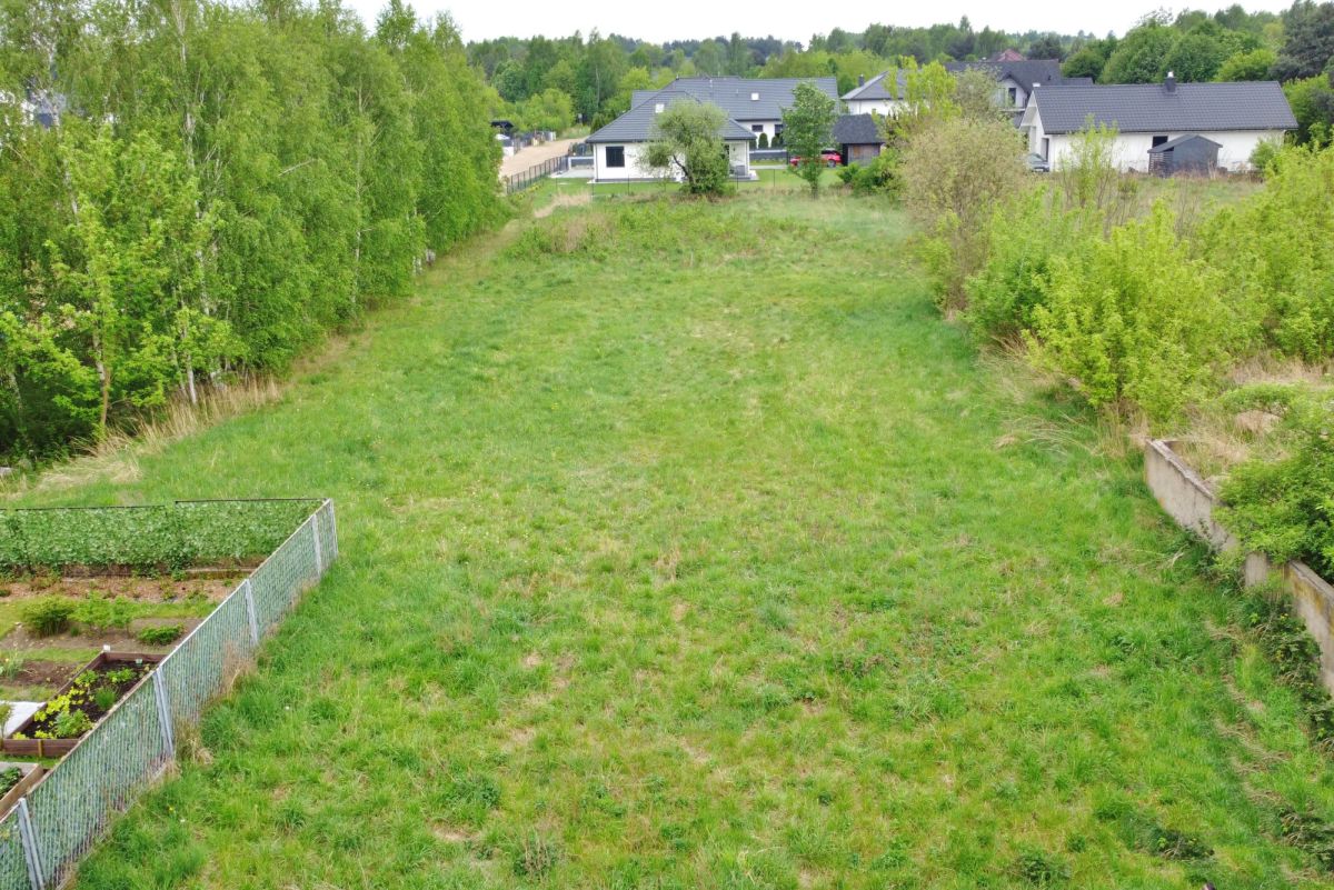 Aerial view of a grassy open plot bordered by trees on both sides, with houses visible in the distance and a fenced garden area in the lower left.