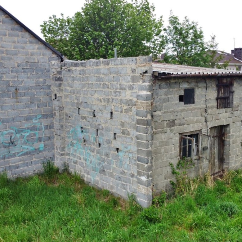 Old gray brick outbuilding with broken windows and a corrugated roof, surrounded by tall grass in a yard