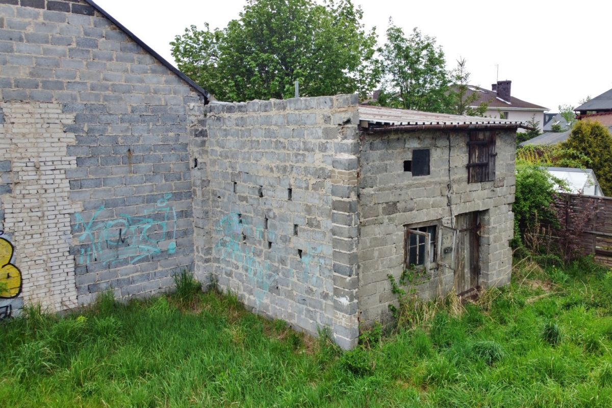 Old gray brick outbuilding with broken windows and a corrugated roof, surrounded by tall grass in a yard