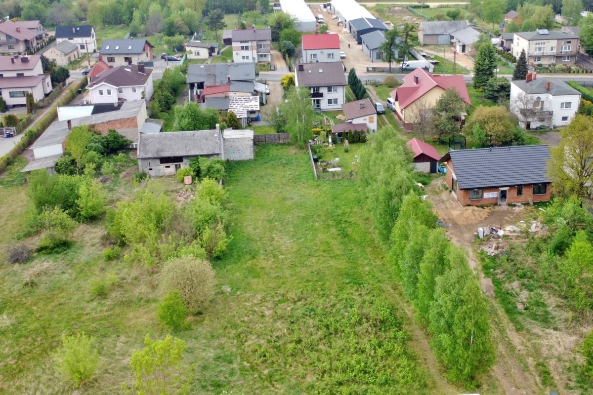 Aerial view of a rural neighborhood with scattered houses, gardens, and a large grassy open lot bordered by trees.