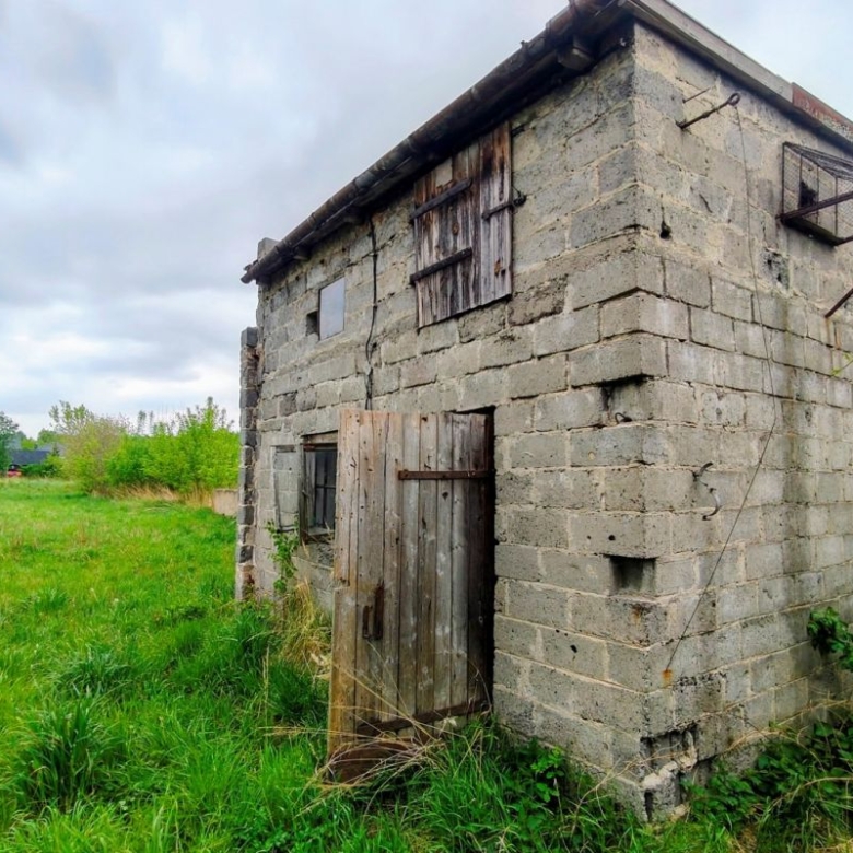 Old concrete outbuilding with a weathered wooden door in a grassy field under a cloudy sky.
