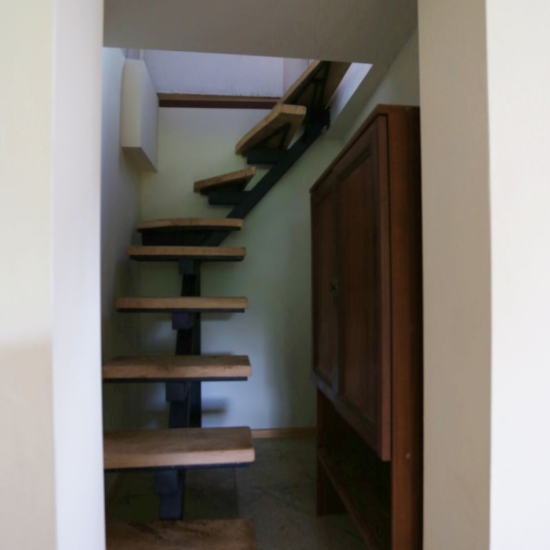 Interior view of a narrow stairway with floating wooden steps and a black metal support, beside a tall wooden cabinet on the right.