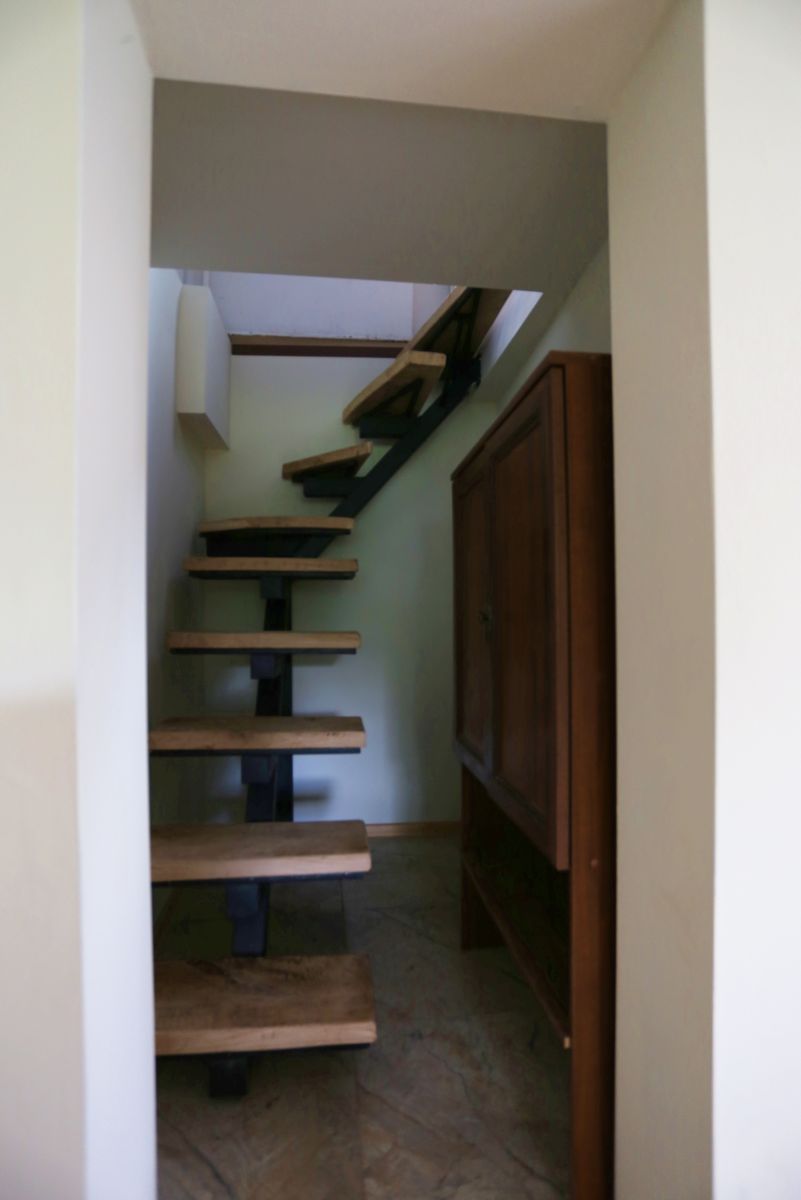 Interior view of a narrow stairway with floating wooden steps and a black metal support, beside a tall wooden cabinet on the right.