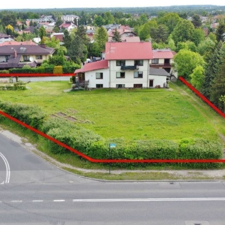 Rectangular grassy property boundary outlined in red along a residential street, with a two-story white house near its center and green trees surrounding it.