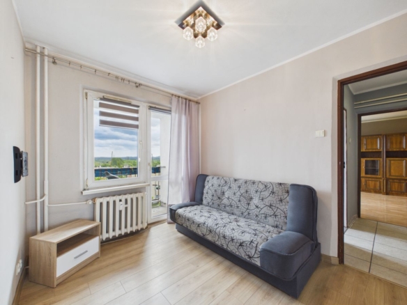 Cozy living room with a patterned gray sofa, wooden floor, and large window/door to a balcony on the left.