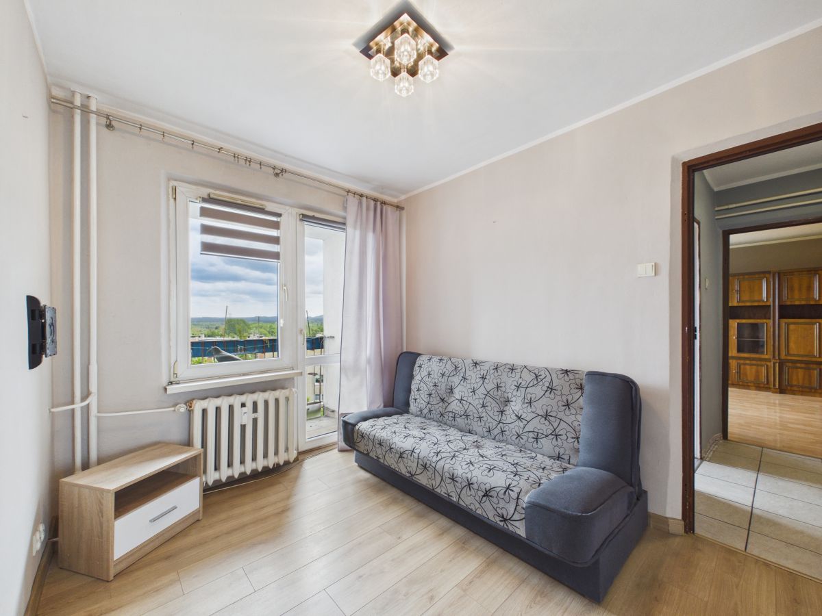 108070862.jpg Cozy living room with a patterned gray sofa, wooden floor, and large window/door to a balcony on the left.