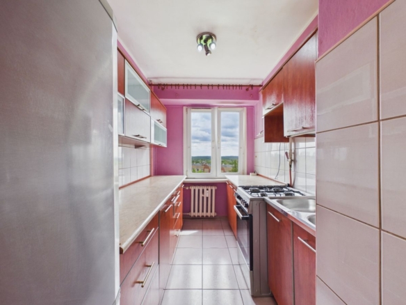 Narrow pink kitchen with wooden cabinets, white countertops, and a window at the far end; fridge on the left, stove and sink along the right side.