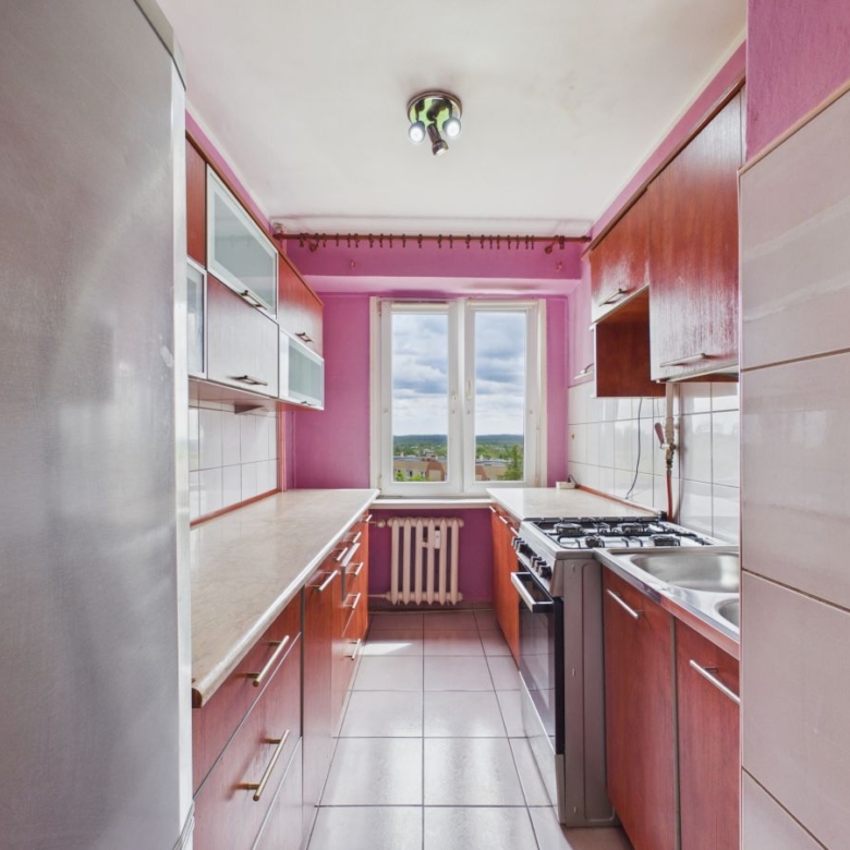 Narrow pink kitchen with wooden cabinets, white countertops, and a window at the far end; fridge on the left, stove and sink along the right side.