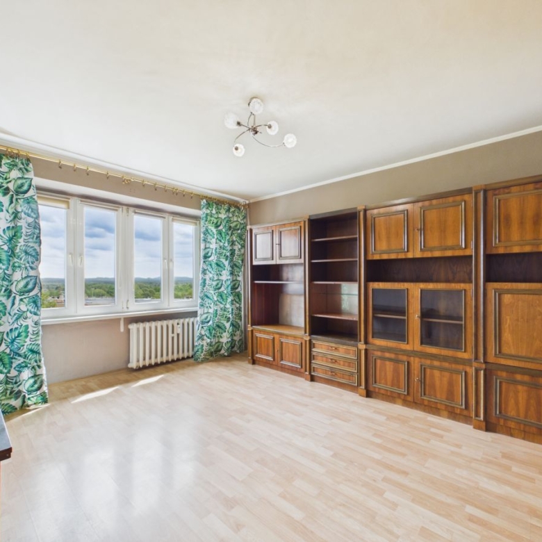 Spacious living room with a large wooden wall unit and cream walls, patterned green leaf curtains by tall windows, and a radiator beneath the view.