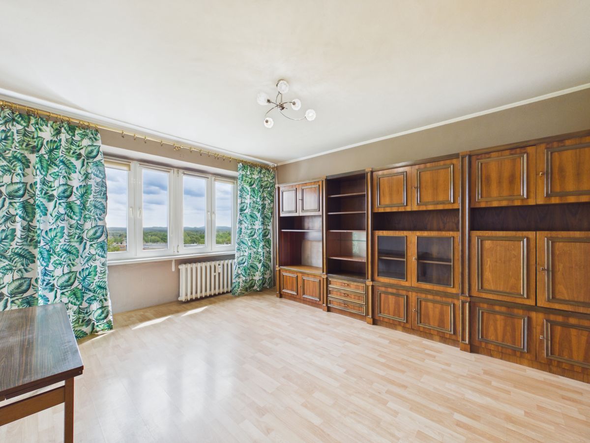 Spacious living room with a large wooden wall unit and cream walls, patterned green leaf curtains by tall windows, and a radiator beneath the view.