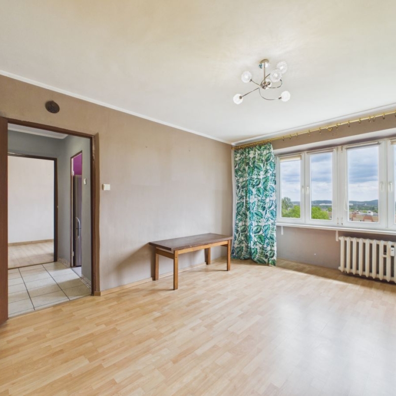 Bright empty living room with light wood flooring, a wooden bench, large windows with green leafy curtains, and a radiator under the windows.