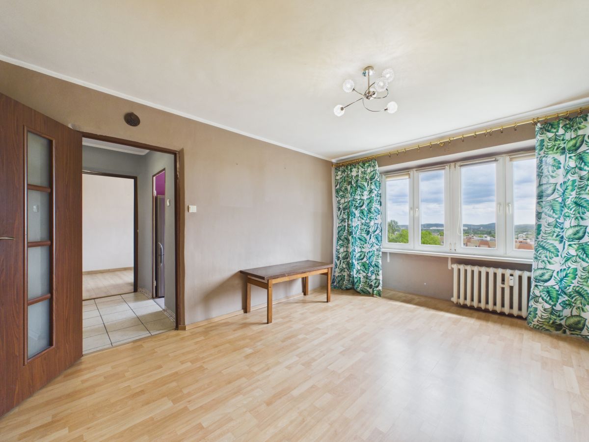 Bright empty living room with light wood flooring, a wooden bench, large windows with green leafy curtains, and a radiator under the windows.