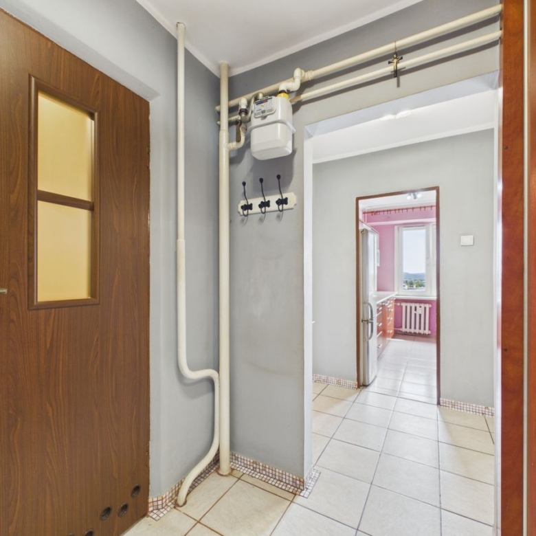Interior hallway with exposed gas meter and white pipes along gray walls, leading to a bright kitchen visible through a doorway on the right side of the frame.