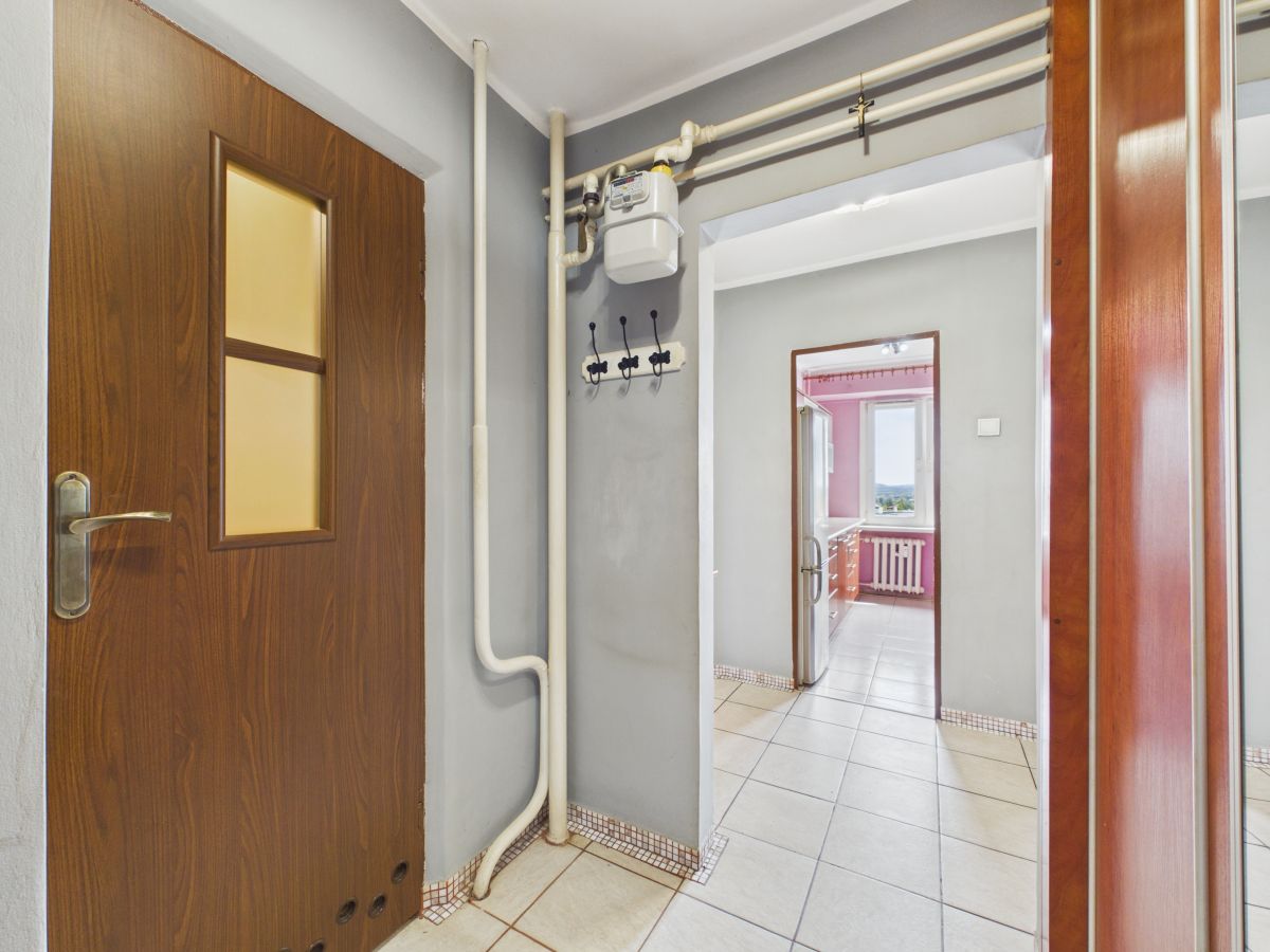 Interior hallway with exposed gas meter and white pipes along gray walls, leading to a bright kitchen visible through a doorway on the right side of the frame.