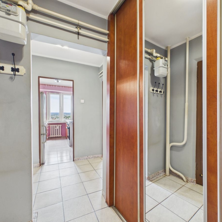 Hallway with gray walls, exposed pipes and gas meter, hooks on the wall, and mirrored wooden closet doors; kitchen visible through the doorway beyond.