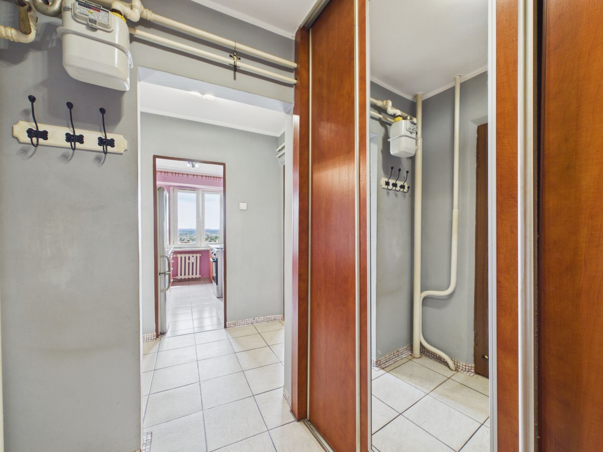 Hallway with gray walls, exposed pipes and gas meter, hooks on the wall, and mirrored wooden closet doors; kitchen visible through the doorway beyond.