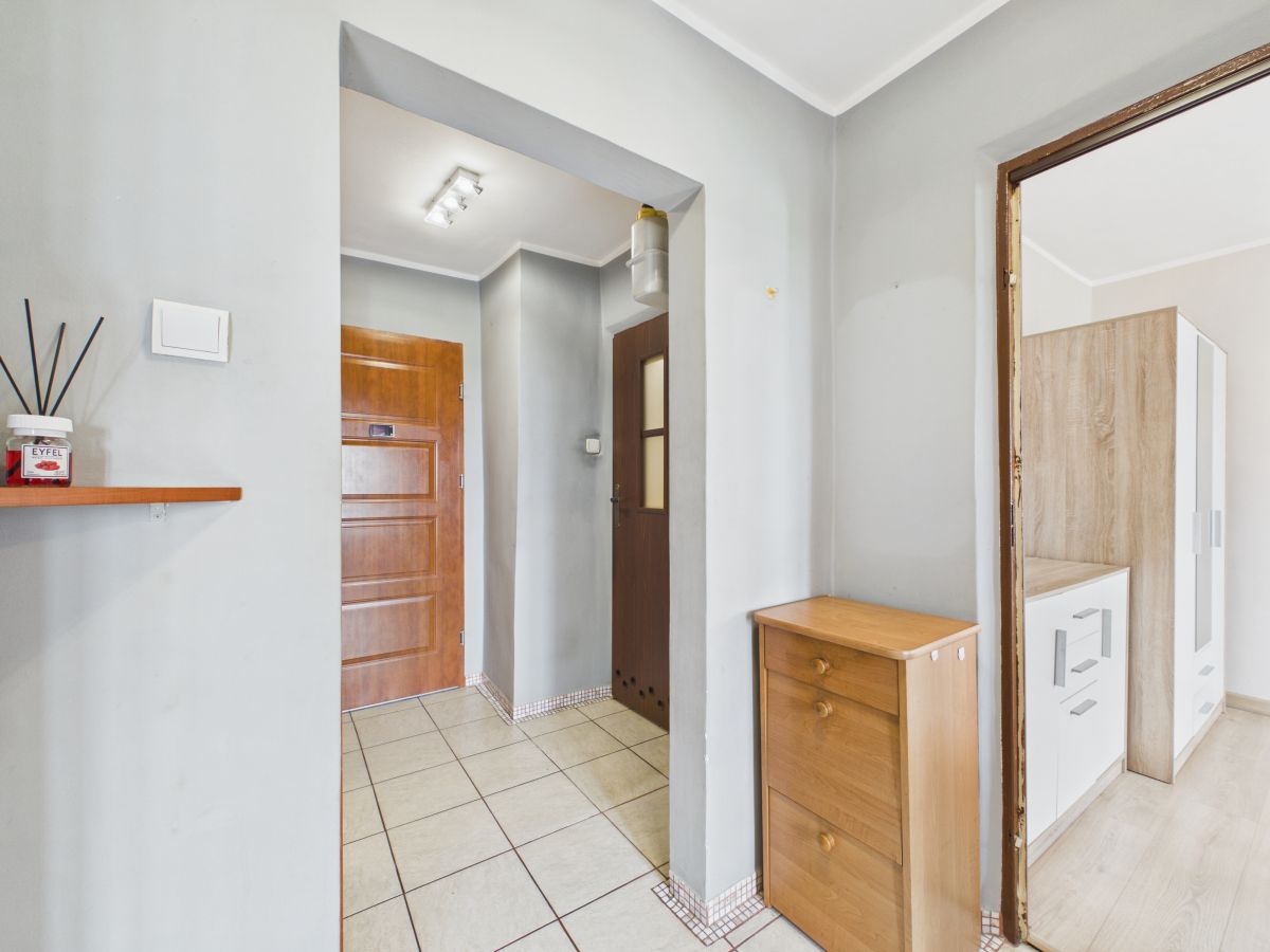 Hallway in a home: gray walls, tiled floor, a wooden front door, and a small three-drawer wooden chest near an opening.