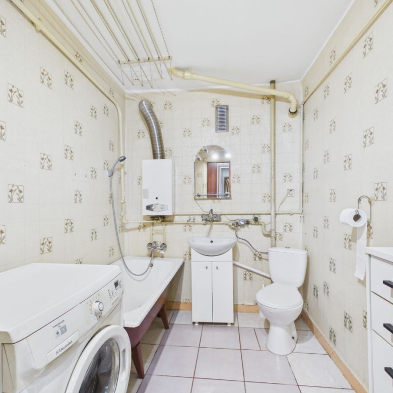 Small bathroom with a washing machine beside a bathtub, sink cabinet, and toilet, exposed pipes along the tiled walls.