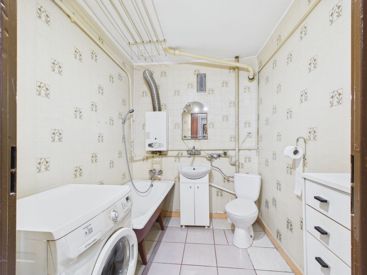 Small bathroom with a washing machine beside a bathtub, sink cabinet, and toilet, exposed pipes along the tiled walls.